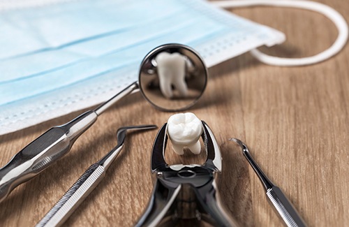 Extracted tooth with forceps, mirror, and other dental instruments next to a mask on a table